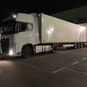 Stovintis sunkvežimis prie iškrovimo platformos, nakties metu su įjungtomis šviesomis.